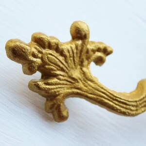 May include: Gold-toned metal drawer pull with a decorative, ornate design. The pull has a curved shape and a detailed, floral-like pattern.