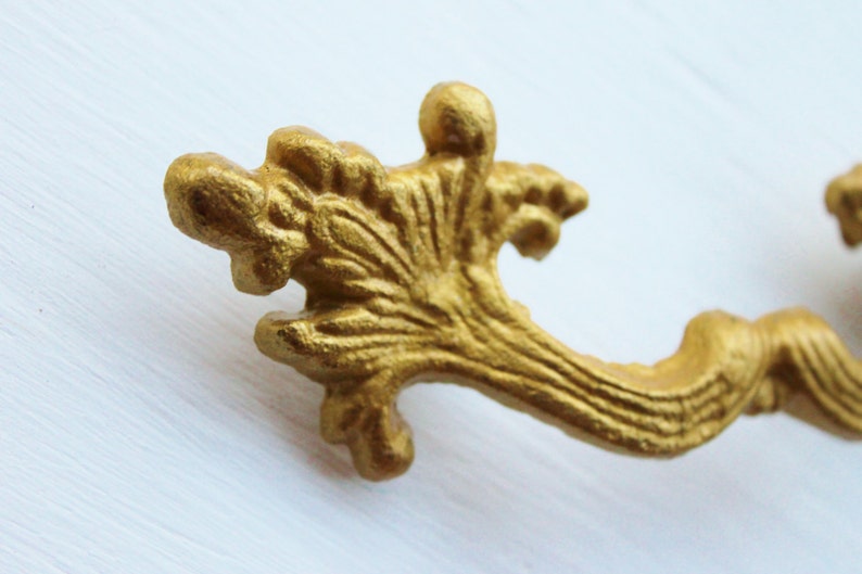 May include: Gold-toned metal drawer pull with a decorative, ornate design. The pull has a curved shape and a detailed, floral-like pattern.