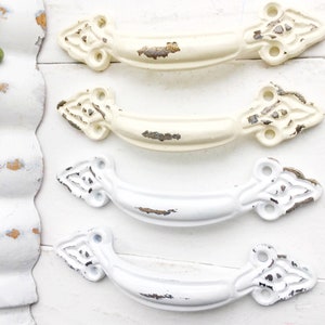 May include: Four antique-style drawer pulls in white and off-white with a distressed finish. The pulls have a decorative, ornate design with a curved shape.