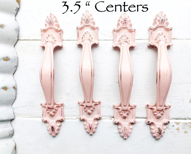 May include: Four pink metal cabinet knobs with ornate detailing. The knobs are 3.5 inches apart.