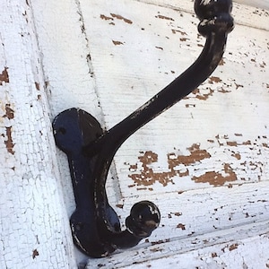 May include: Black metal hook with a rounded top and a single hook. The hook is mounted on a white, weathered wooden surface.
