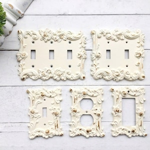 May include: Off-white decorative light switch covers with ornate floral and scrollwork designs. The set includes covers for single, double, and triple switches, a duplex outlet, and a blank plate. Gold accents highlight the details.