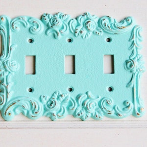 May include: A light blue decorative light switch cover with a floral design. The cover has three switches and is made of metal.