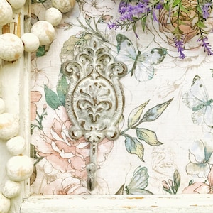 May include: A white metal hook with an ornate design. The hook is against a floral patterned background with butterflies.