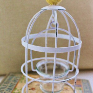 May include: White metal birdcage with a yellow bird on top. The cage has a glass votive holder inside.