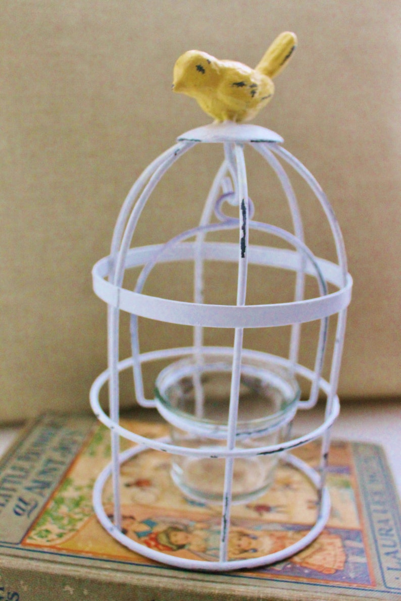 May include: White metal birdcage with a yellow bird on top. The cage has a glass votive holder inside.