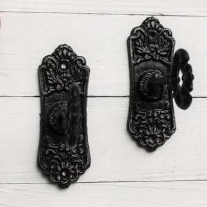 May include: Two black cast iron wall hooks with ornate designs. Each hook features a scroll-like design with a single hook extending from the center.