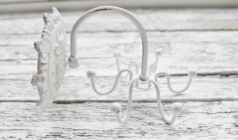May include: White metal wall mounted hook with a decorative scroll design. The hook has five arms that curve out and up to form a hook shape. The hook is mounted on a white wall.