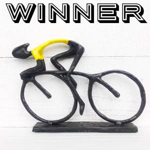 May include: A black metal sculpture of a cyclist in a yellow jersey riding a bicycle. The sculpture is on a black base and the word "WINNER" is written above the sculpture.