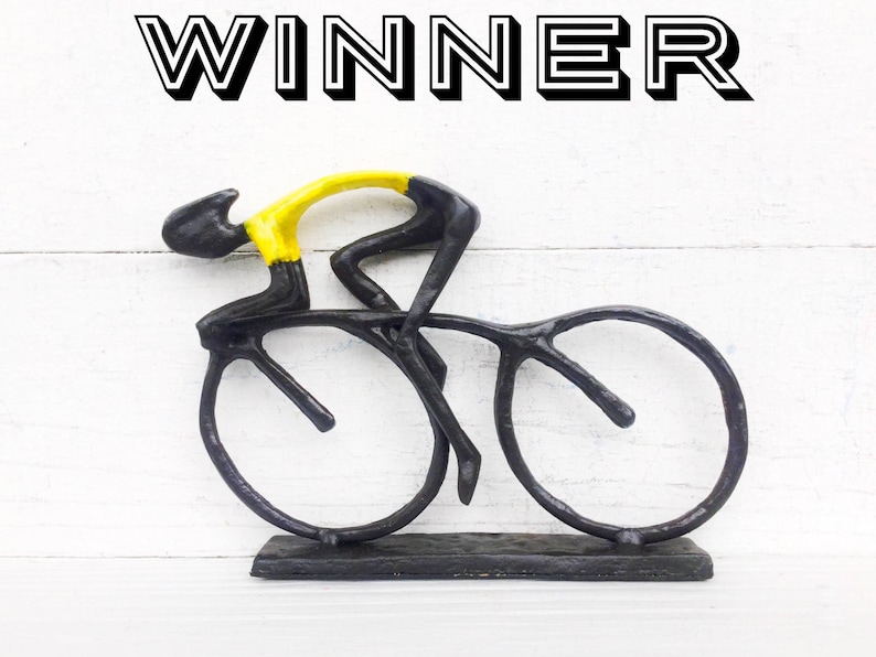 May include: A black metal sculpture of a cyclist in a yellow jersey riding a bicycle. The sculpture is on a black base and the word "WINNER" is written above the sculpture.