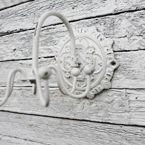May include: White metal wall hook with a decorative floral design. The hook has three prongs for hanging items.