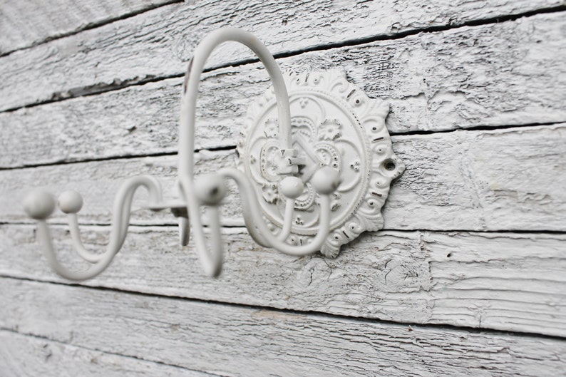 May include: White metal wall hook with a decorative floral design. The hook has three prongs for hanging items.