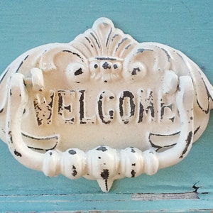 Puede incluir: Un llamador de puerta de metal blanco con un dise&ntilde;o decorativo y la palabra "WELCOME" en letras en relieve.