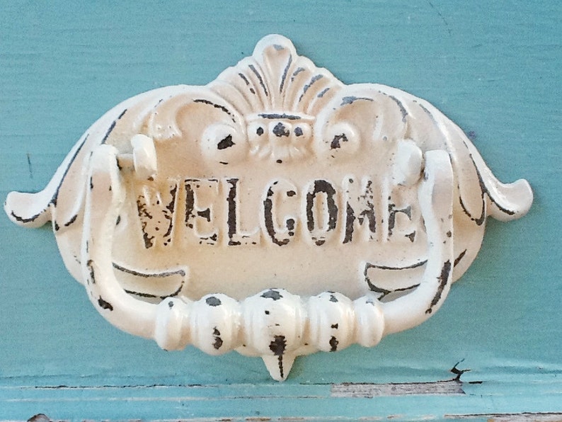 Puede incluir: Un llamador de puerta de metal blanco con un dise&ntilde;o decorativo y la palabra "WELCOME" en letras en relieve.