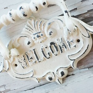 Puede incluir: Un letrero de bienvenida de metal blanco con un asa decorativa. El letrero tiene un acabado desgastado y la palabra "WELCOME" est&aacute; escrita en negro.