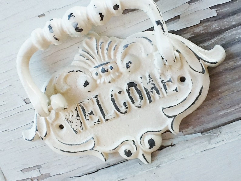 Puede incluir: Un letrero de bienvenida de metal blanco con un asa decorativa. El letrero tiene un acabado desgastado y la palabra "WELCOME" est&aacute; escrita en negro.
