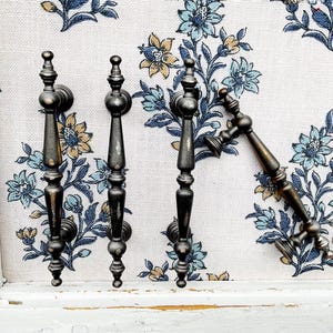 May include: Four antique bronze cabinet pulls with ornate designs. The pulls are arranged on a floral patterned fabric with blue, yellow, and white flowers. The pulls are mounted on a distressed white wooden surface.