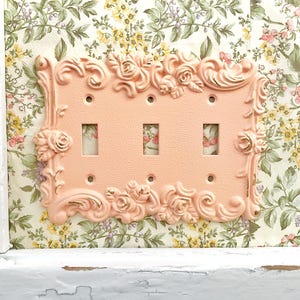 May include: A decorative light switch cover in a soft peach color, adorned with ornate floral and scroll detailing. The cover has three switch openings and is set against a floral wallpaper background. The overall design is vintage-inspired.