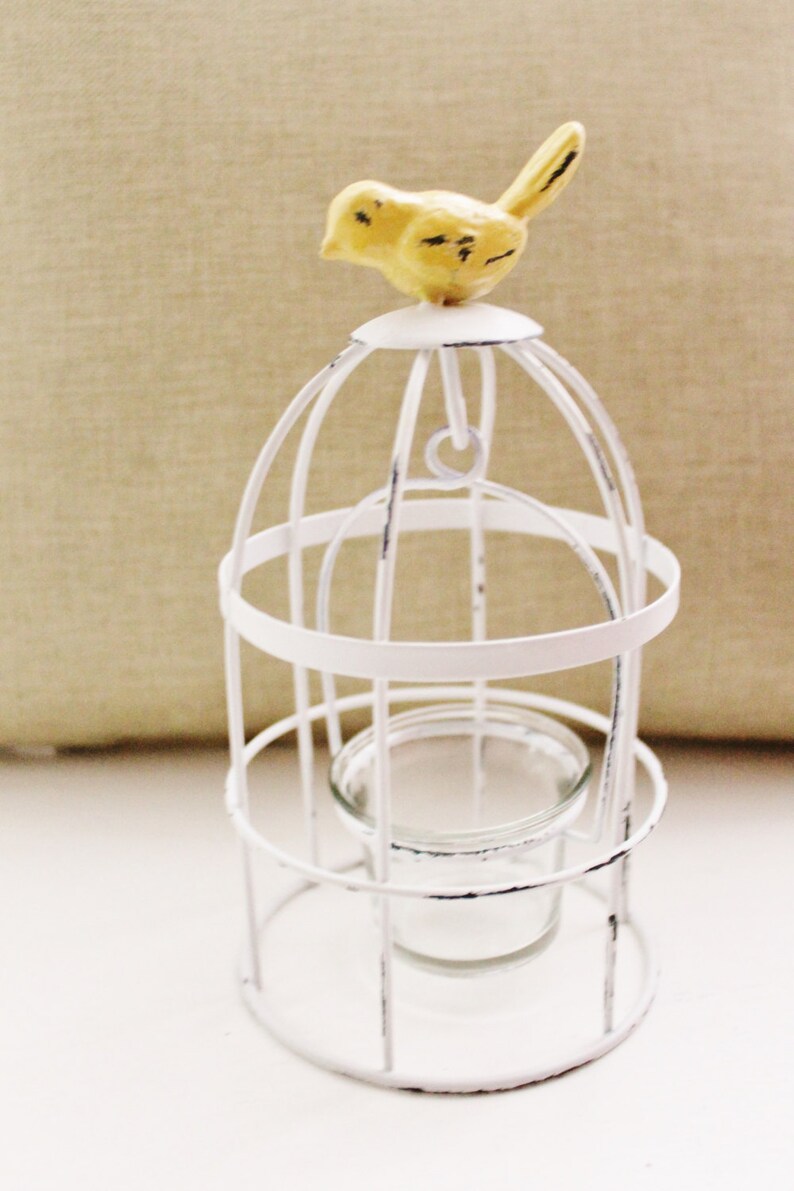 May include: White metal birdcage with a yellow bird figurine on top. The cage has a glass votive holder inside.