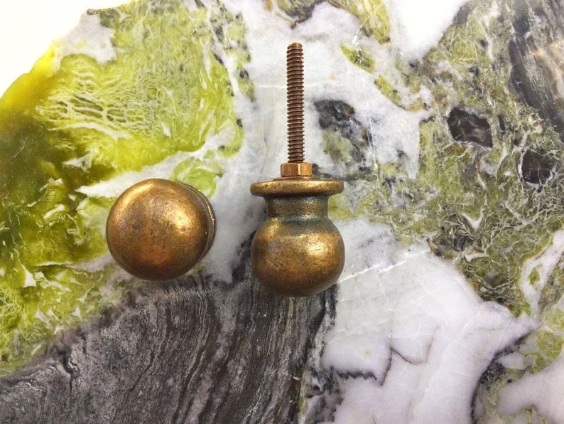 May include: Two antique brass cabinet knobs, one with a threaded stem and the other with a smooth round top. The knobs are displayed on a green and white marble surface.