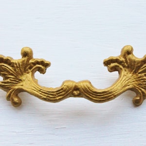 May include: A gold-toned metal drawer pull with a decorative, ornate design. The pull has a curved shape with a floral pattern on each end.