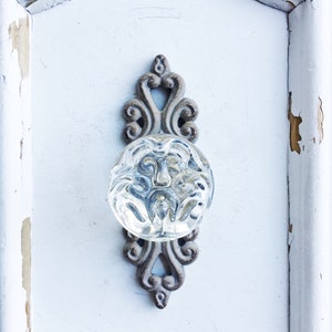 May include: A clear glass doorknob with a decorative, ornate metal backplate. The backplate is a light brown color and has a distressed, vintage look. The doorknob is mounted on a white, distressed wooden surface.