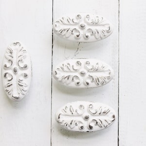 May include: Three white oval-shaped cabinet knobs with an ornate design. The knobs have a distressed white finish and are made of metal.
