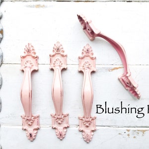 May include: Four ornate pink metal cabinet door pulls with a decorative design. The pulls are arranged in a row on a white wooden surface. The text "Blushing Pink" is written below the pulls.