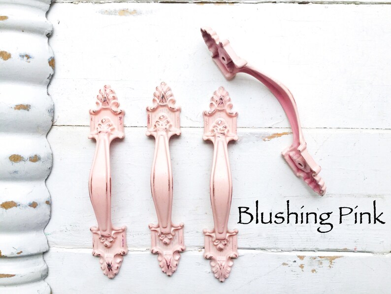 May include: Four ornate pink metal cabinet door pulls with a decorative design. The pulls are arranged in a row on a white wooden surface. The text "Blushing Pink" is written below the pulls.