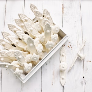 May include: A white box filled with white ceramic drawer pulls with a floral design. The drawer pulls are arranged in a fan shape. There are two additional drawer pulls on a white wooden surface.