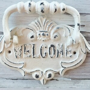 Puede incluir: Un letrero de bienvenida de metal blanco con un asa decorativa. El letrero tiene un acabado desgastado y la palabra "WELCOME" est&aacute; escrita en negro.