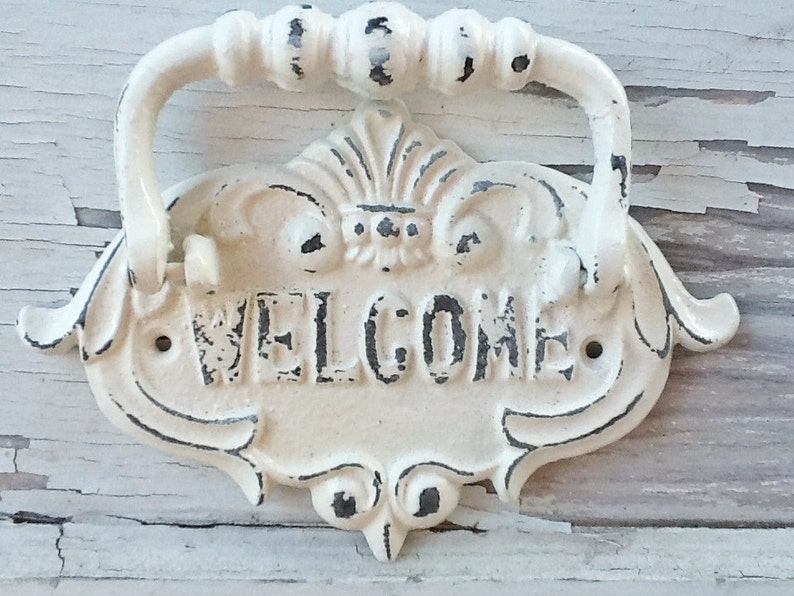 Puede incluir: Un letrero de bienvenida de metal blanco con un asa decorativa. El letrero tiene un acabado desgastado y la palabra "WELCOME" est&aacute; escrita en negro.