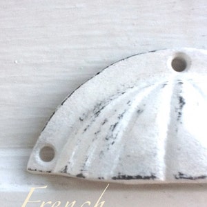 May include: White painted metal cabinet knob with a scalloped design. The knob has two holes for mounting. The text "French inspired..." is written below the knob.