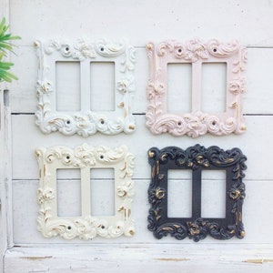 May include: Four decorative light switch covers in different colours and styles. The covers are white, pink, cream, and black with gold accents. Each cover has a double switch.
