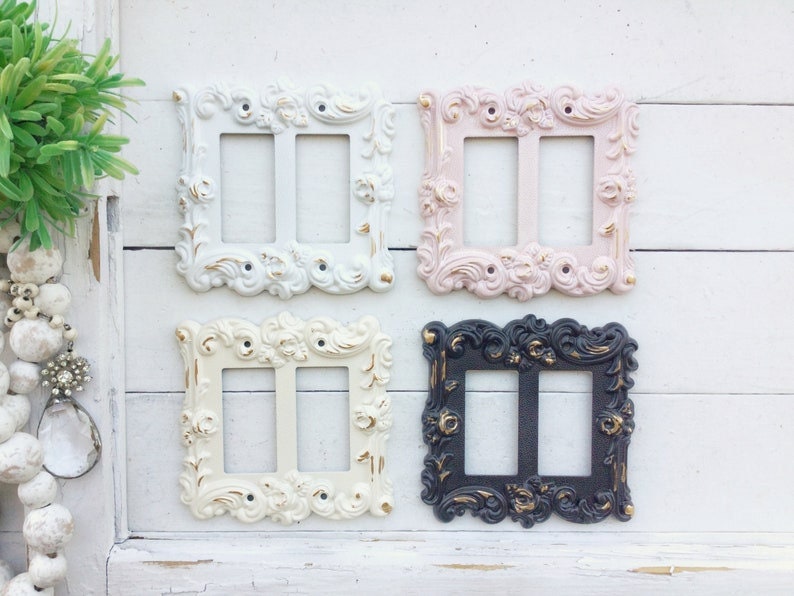 May include: Four decorative light switch covers in different colours and styles. The covers are white, pink, cream, and black with gold accents. Each cover has a double switch.