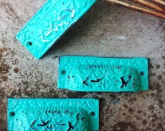May include: Three turquoise blue metal drawer pulls with intricate designs. The pulls are rectangular and have a slightly curved shape.