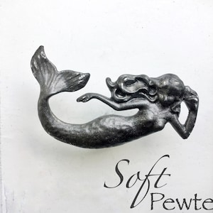 May include: A silver mermaid figurine with flowing hair and a tail. The figurine is on a white background with the words "Soft Pewter" written below it.