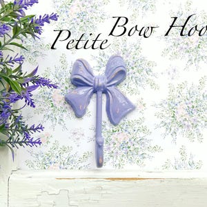 May include: A lavender-colored ceramic bow hook with the text "Petite Bow Hook" on a white background with a floral pattern.