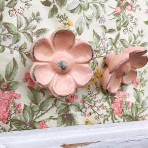 May include: Two pink flower-shaped cabinet knobs with distressed edges. The knobs are set against a floral patterned background with pink, green, and yellow flowers. The knobs are likely made of metal.