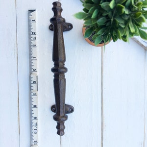 May include: A dark brown cast iron door pull, approximately 13 inches long, with decorative detailing. The handle is mounted on a white wooden surface next to a small potted plant and a measuring tape.