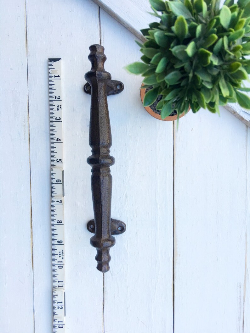 May include: A dark brown cast iron door pull, approximately 13 inches long, with decorative detailing. The handle is mounted on a white wooden surface next to a small potted plant and a measuring tape.
