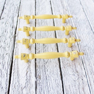 May include: Four yellow metal cabinet knobs with a distressed finish. The knobs are arranged in a row on a white wooden surface.