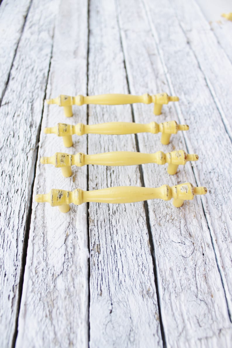 May include: Four yellow metal cabinet knobs with a distressed finish. The knobs are arranged in a row on a white wooden surface.