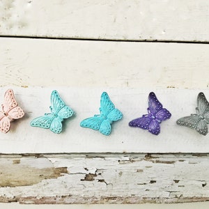 K&ouml;nnte beinhalten: F&uuml;nf dekorative Schmetterlingsornamente in einer Reihe. Die Schmetterlinge sind in den Farbt&ouml;nen Rosa, T&uuml;rkis, T&uuml;rkis, Lila und Grau gehalten. Die Schmetterlinge befinden sich auf einer wei&szlig;en, verwitterten Holzoberfl&auml;che.