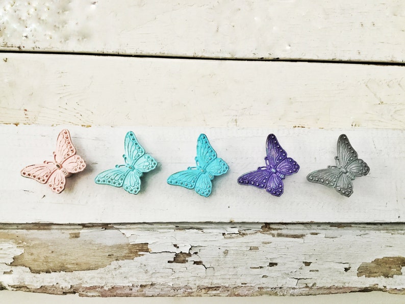 K&ouml;nnte beinhalten: F&uuml;nf dekorative Schmetterlingsornamente in einer Reihe. Die Schmetterlinge sind in den Farbt&ouml;nen Rosa, T&uuml;rkis, T&uuml;rkis, Lila und Grau gehalten. Die Schmetterlinge befinden sich auf einer wei&szlig;en, verwitterten Holzoberfl&auml;che.