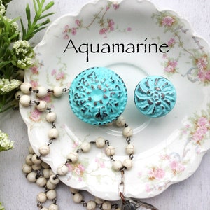 May include: Two aqua blue cabinet knobs with a distressed finish. The knobs have a floral design and are displayed on a white plate with pink floral accents. The text "Aquamarine" is written on the plate.