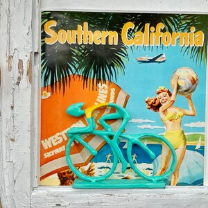 May include: A turquoise blue bicycle figurine with a yellow rider, set against a vintage travel poster for Southern California. The poster features palm trees, a plane, and a woman holding a beach ball.