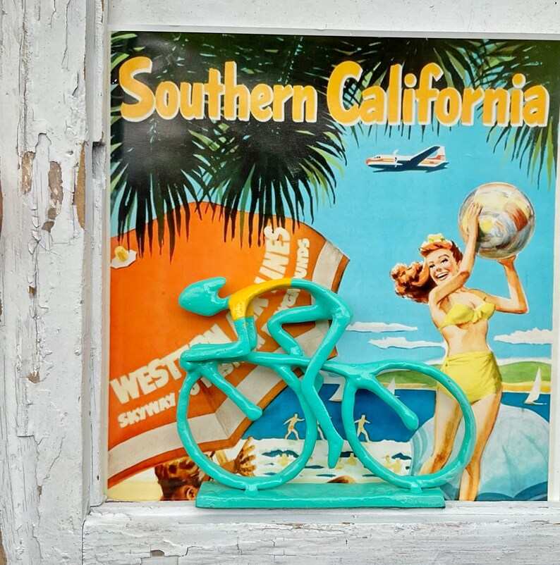 May include: A turquoise blue bicycle figurine with a yellow rider, set against a vintage travel poster for Southern California. The poster features palm trees, a plane, and a woman holding a beach ball.