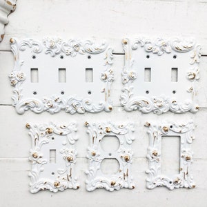 May include: Set of five white decorative light switch covers with ornate floral designs and gold accents. The covers are in various sizes and shapes.
