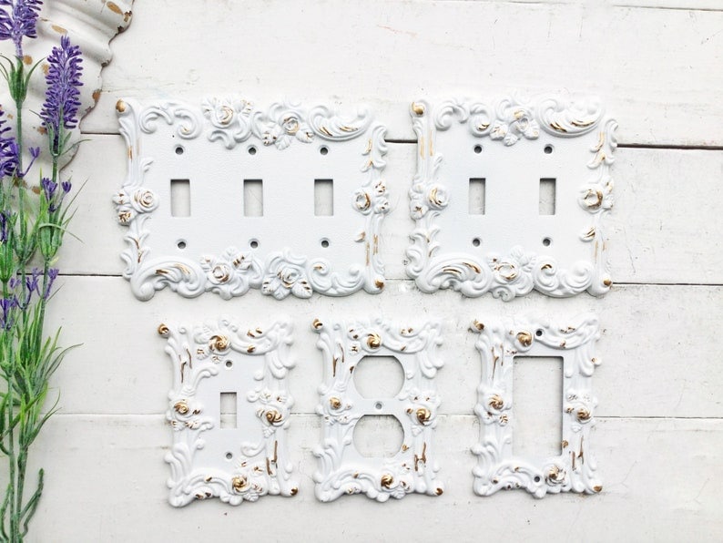May include: Set of five white decorative light switch covers with ornate floral designs and gold accents. The covers are in various sizes and shapes.